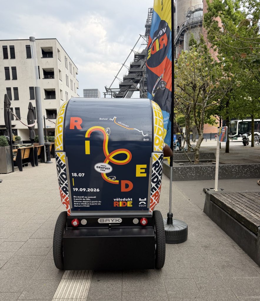 The Vëlodukt Ride bike taxi parked on the Belval esplanade, with the blast furnaces behind.