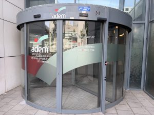 The curved glass entrance of the ADEM employment agency in Belval, with the adem logo and a panel listing opening hours of 8.00 to 12.00 and 13.00 to 17.00, Monday to Friday.