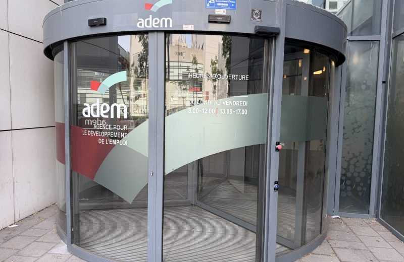 The curved glass entrance of the ADEM employment agency in Belval, with the adem logo and a panel listing opening hours of 8.00 to 12.00 and 13.00 to 17.00, Monday to Friday.