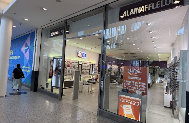 The Alain Afflelou shopfront at Belval Plaza (shown mirrored in the photograph, shot through glass), with the Afflelou wordmark above the entrance and a blue promotional wall panel reading "Belval Plaza 2" visible behind the display.