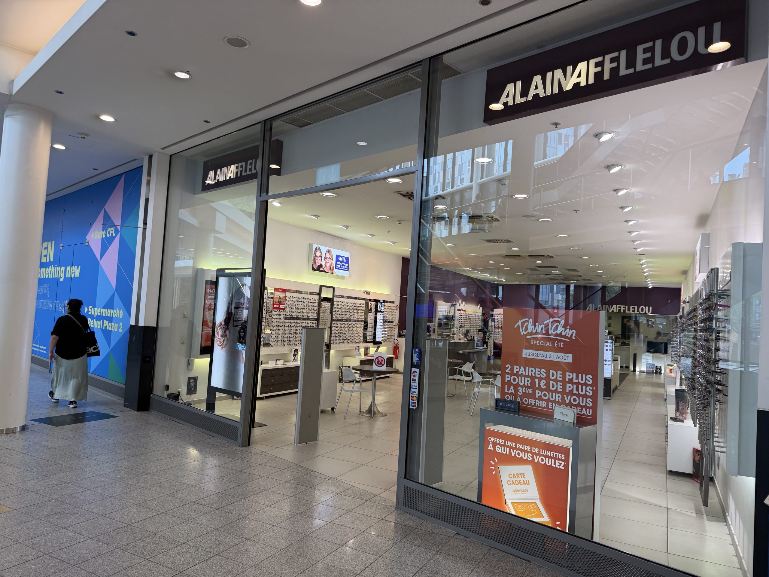 The Alain Afflelou shopfront at Belval Plaza (shown mirrored in the photograph, shot through glass), with the Afflelou wordmark above the entrance and a blue promotional wall panel reading "Belval Plaza 2" visible behind the display.