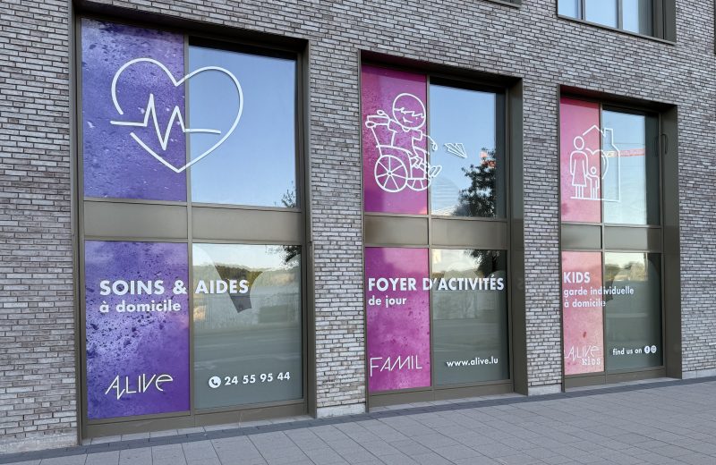 Three shopfront windows for Alive in Belval, each with a different coloured panel: a purple panel with a heart and heartbeat line reading "Soins & Aides à domicile" and the Alive logo and phone number, a magenta panel with a child in a wheelchair reading "Foyer d'Activités de jour" and the FAMIL name, and a pink panel with a parent and child icon reading "Kids, garde individuelle à domicile" and the Alive Kids name.