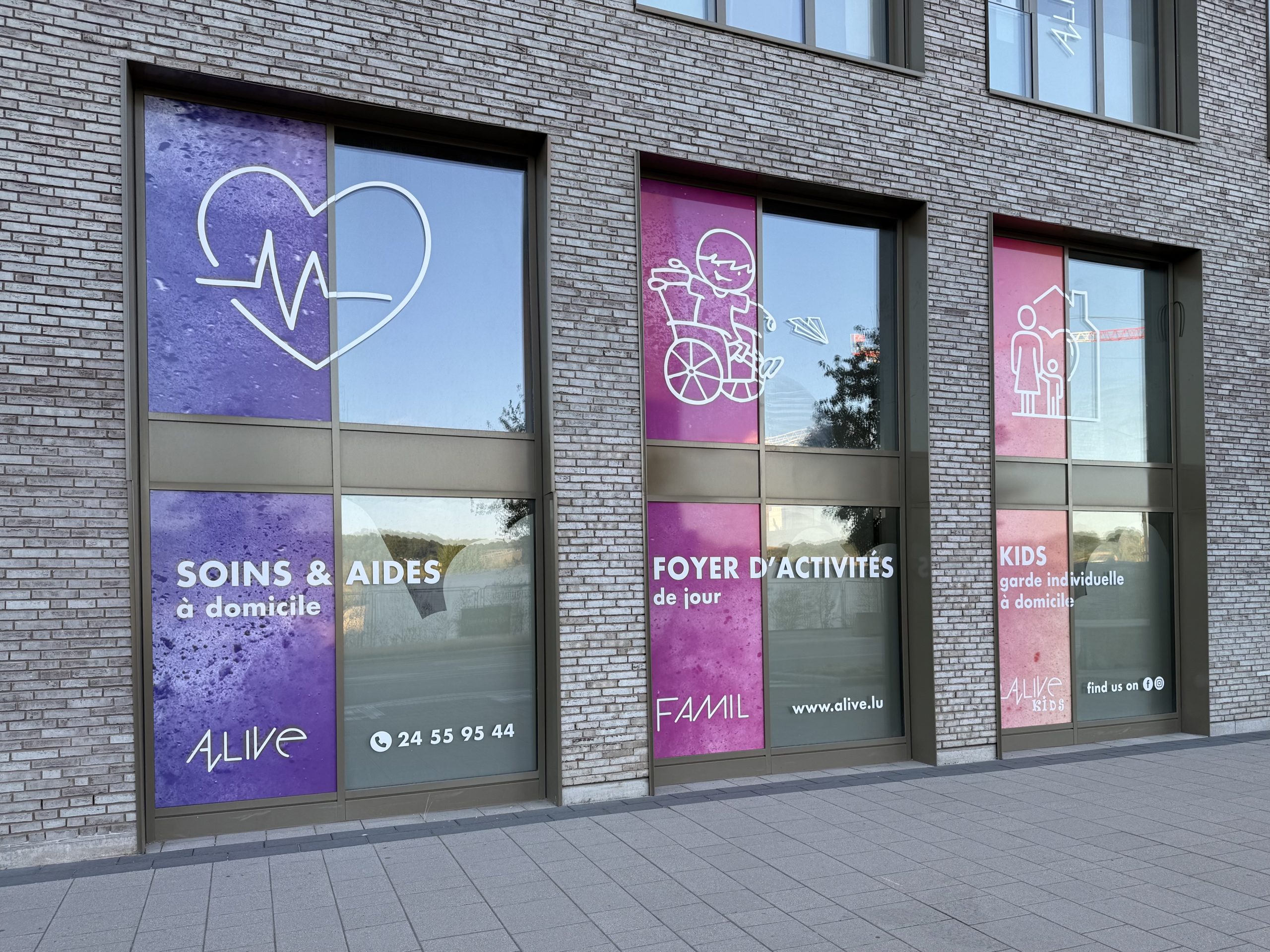 Three shopfront windows for Alive in Belval, each with a different coloured panel: a purple panel with a heart and heartbeat line reading "Soins & Aides à domicile" and the Alive logo and phone number, a magenta panel with a child in a wheelchair reading "Foyer d'Activités de jour" and the FAMIL name, and a pink panel with a parent and child icon reading "Kids, garde individuelle à domicile" and the Alive Kids name.