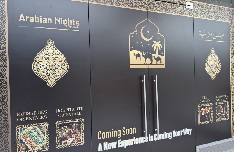 The black and gold hoarding around the future Arabian Nights unit in Belval Plaza II, with the Arabian Nights by Jnede name in gold, a camel and crescent moon motif over the doors, photographs of baklava and Turkish delight, and the words Coming Soon, A New Experience is Coming Your Way.