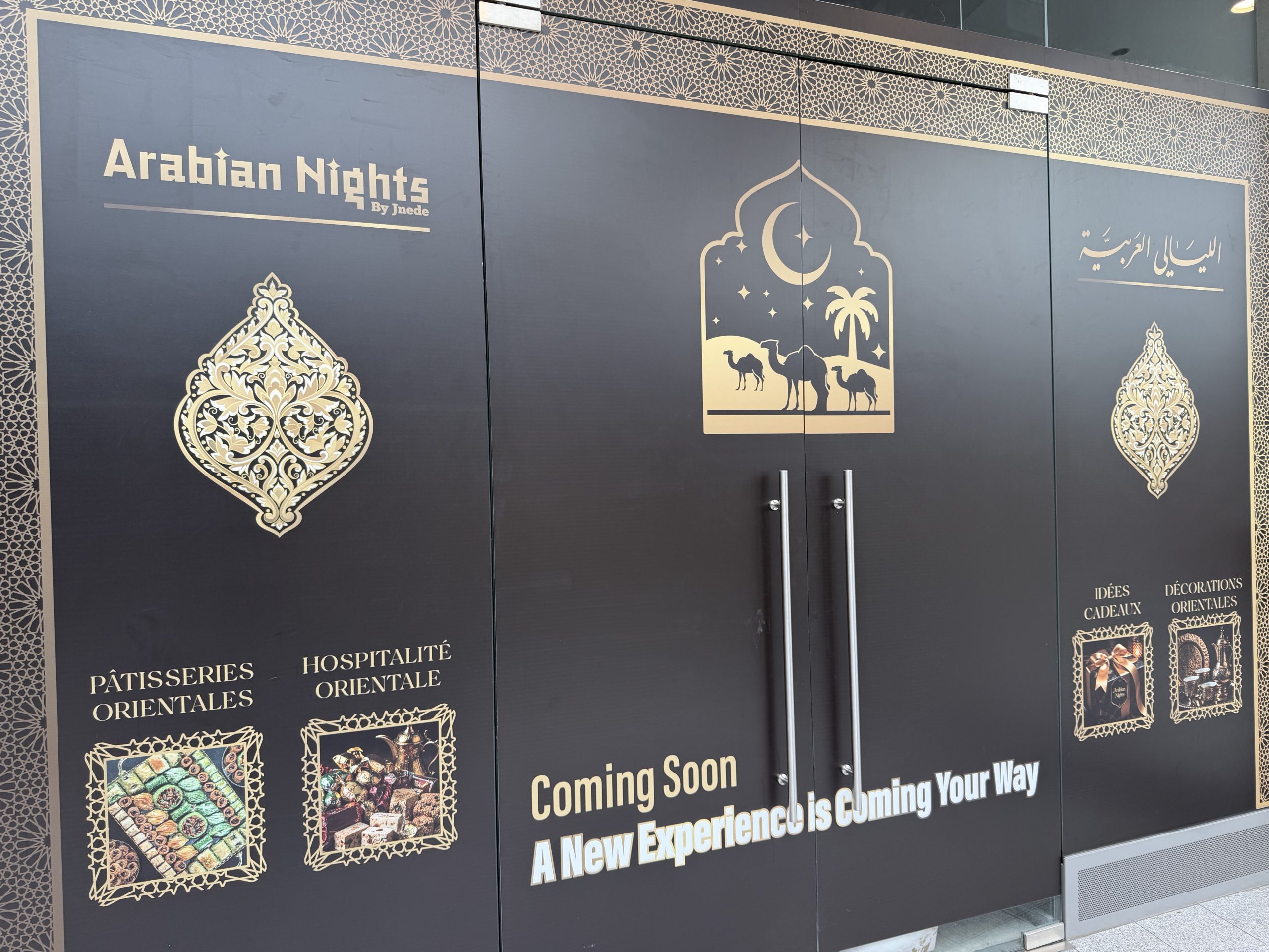 The black and gold hoarding around the future Arabian Nights unit in Belval Plaza II, with the Arabian Nights by Jnede name in gold, a camel and crescent moon motif over the doors, photographs of baklava and Turkish delight, and the words Coming Soon, A New Experience is Coming Your Way.