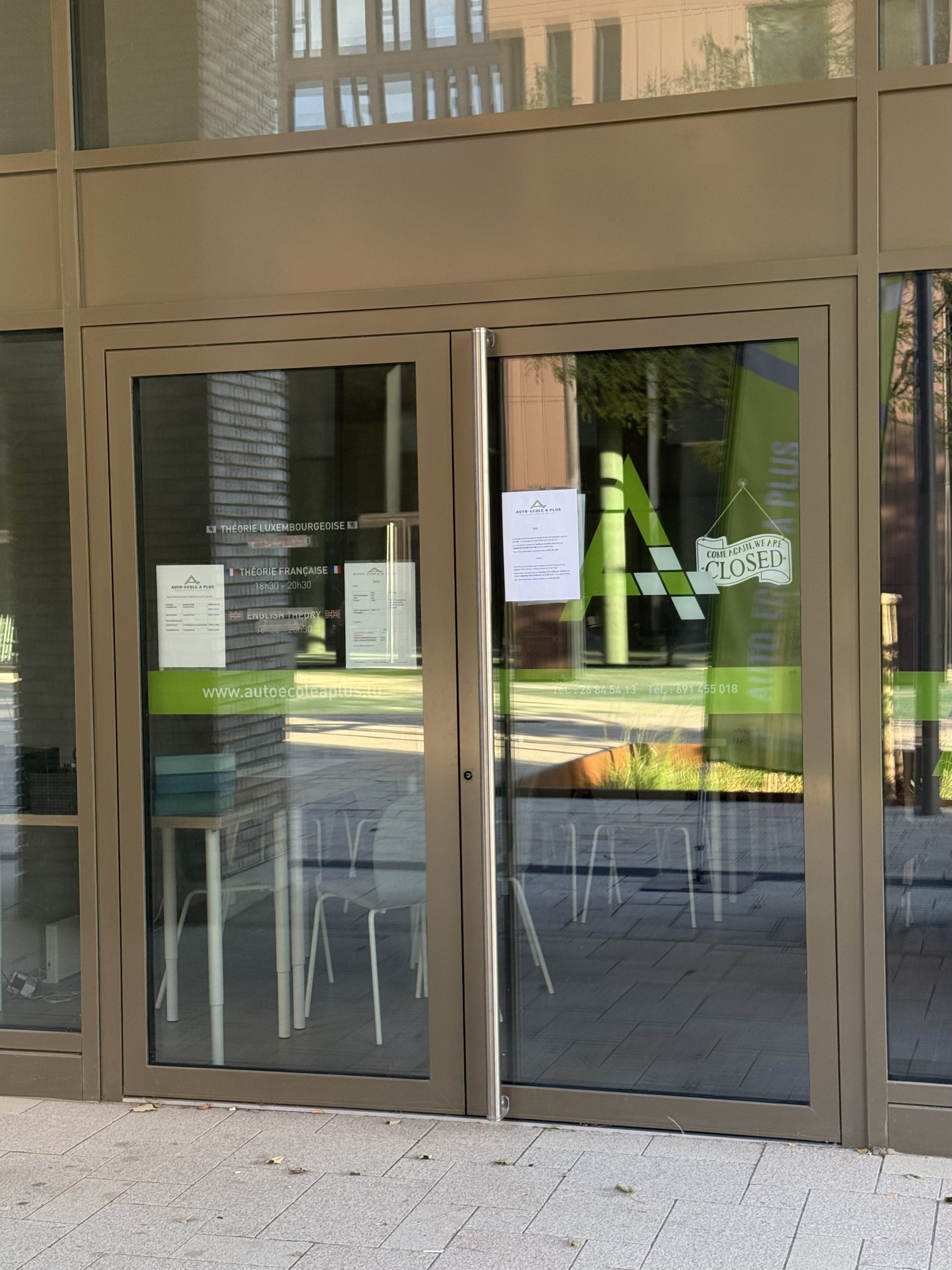 The glass entrance of Auto-École A Plus in Belval, with the green A Plus logo, a closed sign and a notice taped to the door listing theory class times in French, Luxembourgish and English.