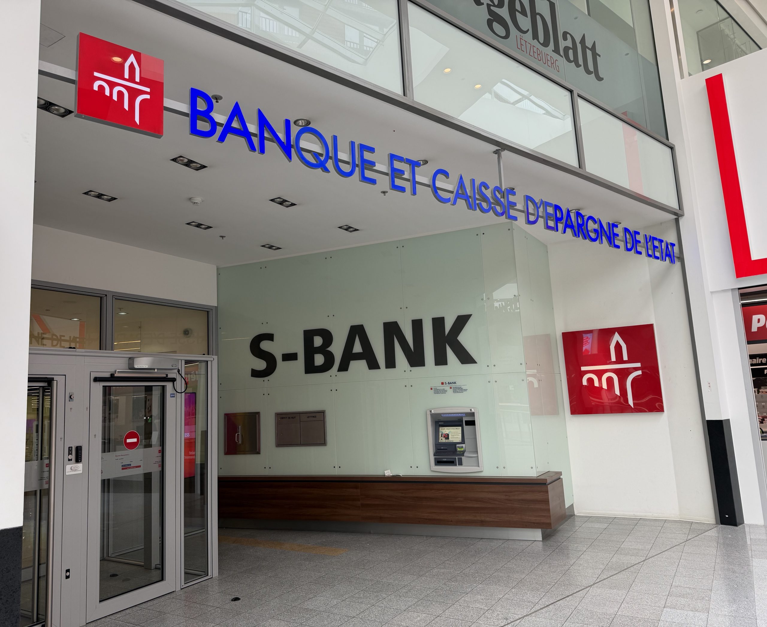 The S-BANK and BCEE branded frontage of the Spuerkeess bank branch at Belval Plaza (shown mirrored in the photograph, shot through glass), with the Spuerkeess wordmark above the entrance.