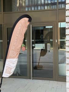A promotional flag outside Beauty Palace reading "Beauty Palace, Esthetic & Wellness" with separate phone numbers listed for the Lamadelaine and Belval branches, a QR code, and a 2025 Top Rated on Salonkee five-star badge, with a price list for pedicure and body treatments visible through the glass door behind it.
