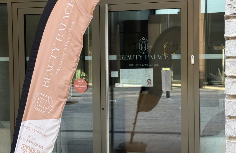 A promotional flag outside Beauty Palace reading "Beauty Palace, Esthetic & Wellness" with separate phone numbers listed for the Lamadelaine and Belval branches, a QR code, and a 2025 Top Rated on Salonkee five-star badge, with a price list for pedicure and body treatments visible through the glass door behind it.