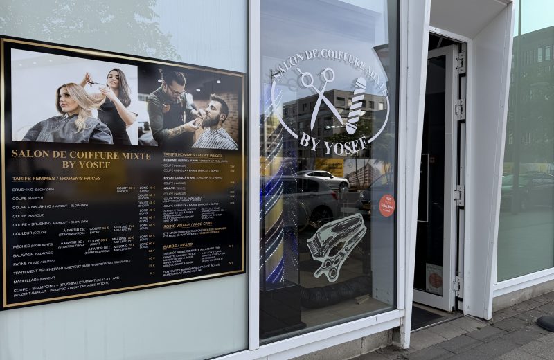 The By Yosef salon in Belval, with a large price board in French and English in the window and the salon logo etched on the glass.
