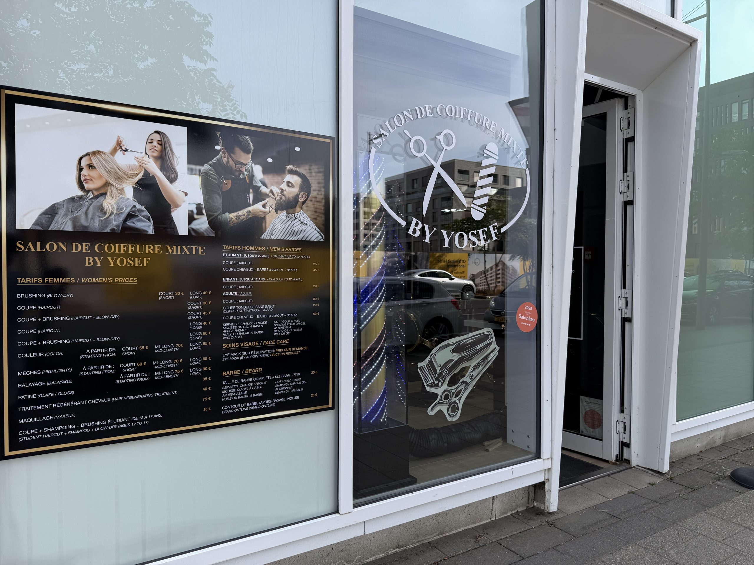 The By Yosef salon in Belval, with a large price board in French and English in the window and the salon logo etched on the glass.