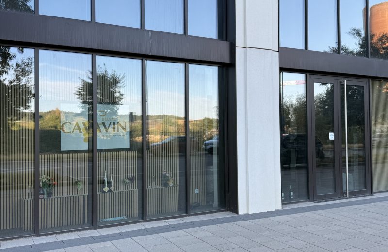 The Cavavin Belval shopfront, a glass-fronted unit with CAVAVIN BELVAL and the line "la passion du conseil" printed on the window, bottles displayed on shelving inside, and the reflection of an orange blast furnace tower and a red construction crane visible in the glass.