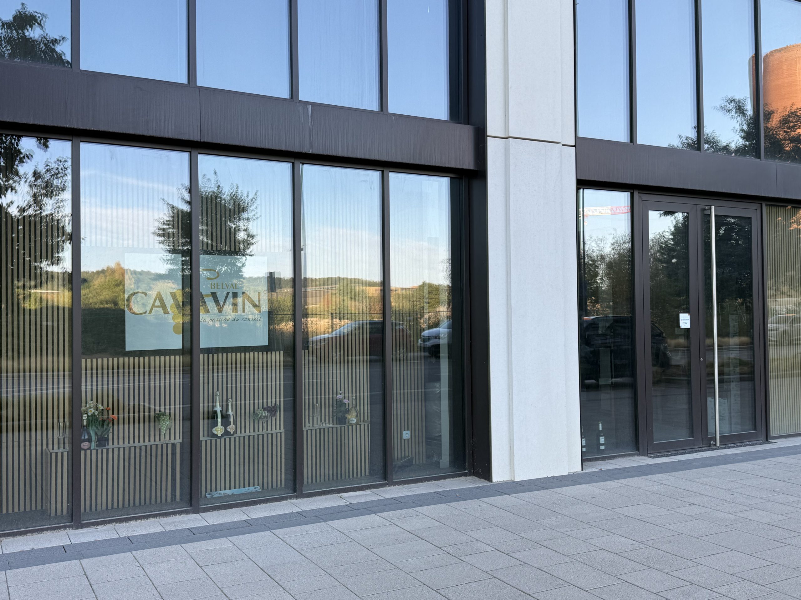 The Cavavin Belval shopfront, a glass-fronted unit with CAVAVIN BELVAL and the line "la passion du conseil" printed on the window, bottles displayed on shelving inside, and the reflection of an orange blast furnace tower and a red construction crane visible in the glass.