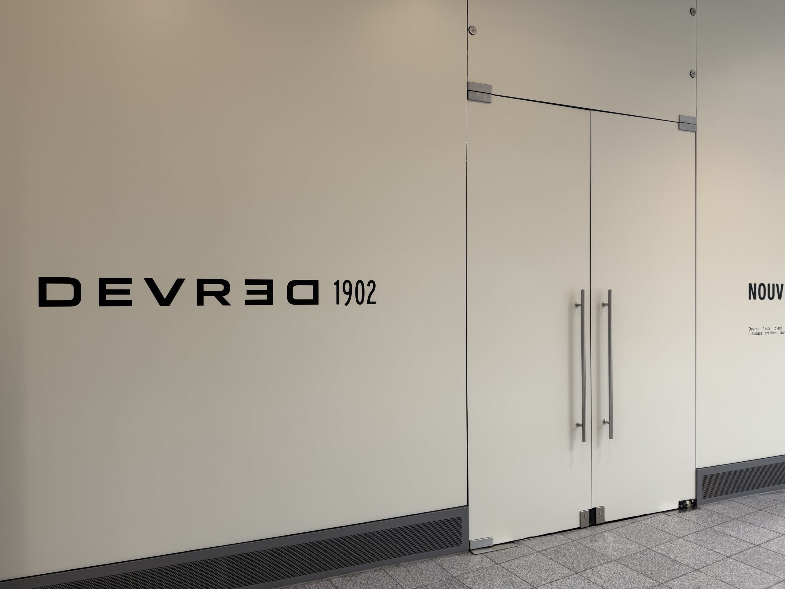 The hoarding around the future Devred 1902 unit in Belval Plaza II, a pale panel carrying the black DEVRED 1902 wordmark beside glass doors, with the brand's own text panel to the right.