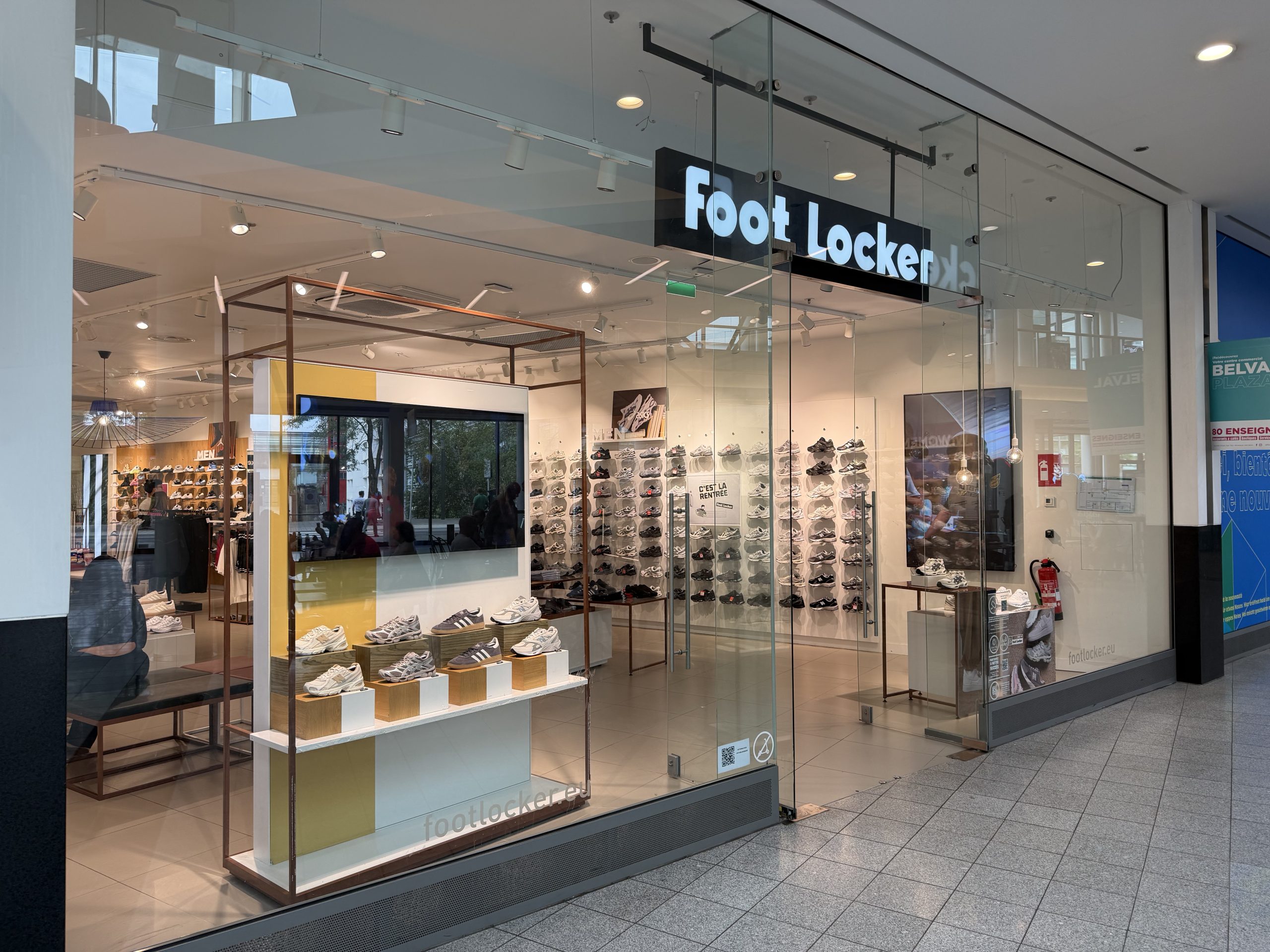 The Foot Locker shopfront in Belval Plaza, with the black Foot Locker sign above the entrance and rows of trainers displayed on the wall inside, footlocker.eu printed on the glass.