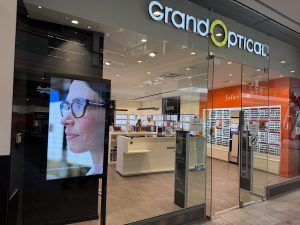 The GrandOptical shopfront in Belval Plaza, with the orange Solaris sunglasses section visible inside and a display screen beside the entrance.