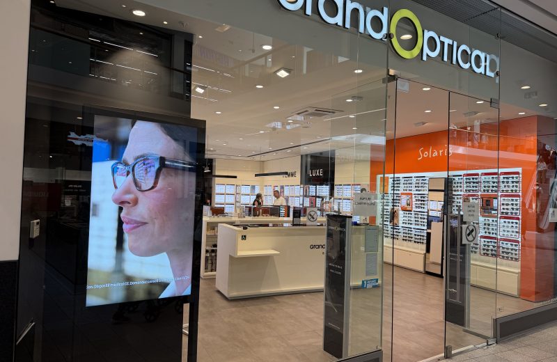 The GrandOptical shopfront in Belval Plaza, with the orange Solaris sunglasses section visible inside and a display screen beside the entrance.