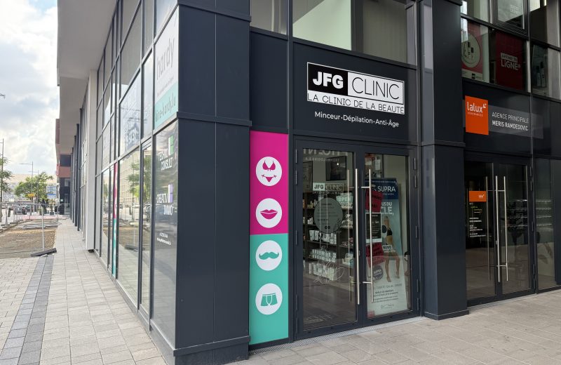 The JFG Clinic Belval shopfront on boulevard du Jazz, dark fascia with a black and white "JFG CLINIC" sign reading "La Clinic de la Beauté" and "Minceur, Dépilation, Anti-Âge" beneath it, a tall pink and teal panel beside the door with four white icons for a bikini top, lips, a moustache and underwear, and glass doors showing a printed hours poster, a phone number and promotional posters for the SUPRA body sculpting system inside.
