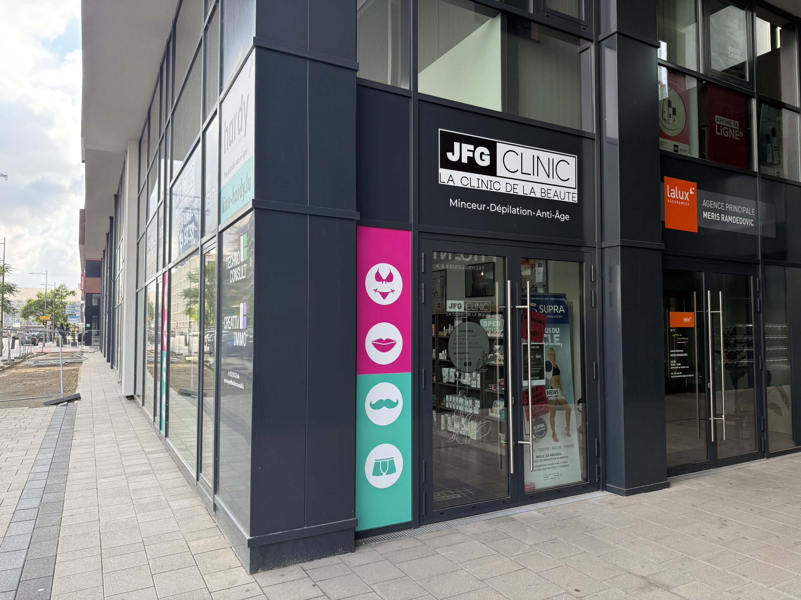 The JFG Clinic Belval shopfront on boulevard du Jazz, dark fascia with a black and white "JFG CLINIC" sign reading "La Clinic de la Beauté" and "Minceur, Dépilation, Anti-Âge" beneath it, a tall pink and teal panel beside the door with four white icons for a bikini top, lips, a moustache and underwear, and glass doors showing a printed hours poster, a phone number and promotional posters for the SUPRA body sculpting system inside.