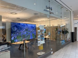 Jerry Travel shopfront at Belval Plaza, glass frontage with the Jerry Travel wordmark and wing logo above the door, a large screen inside playing aerial footage of Madeira, printed holiday brochures on a display stand, and a shelf of model aircraft near the window.