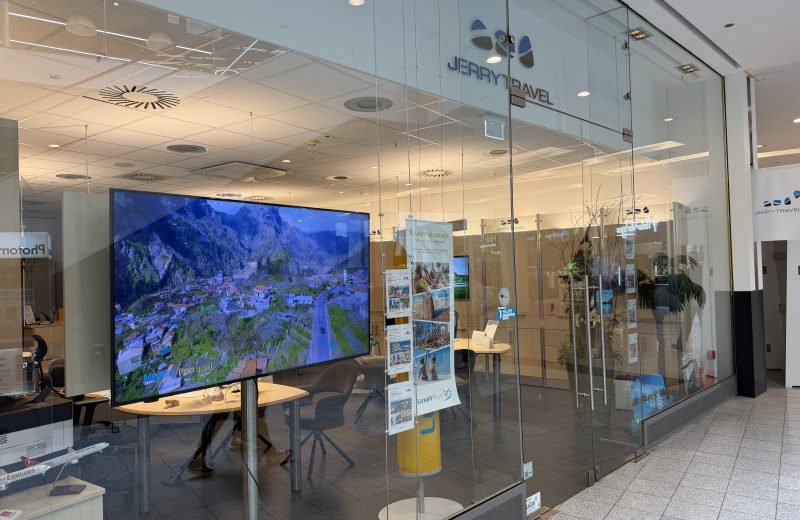 Jerry Travel shopfront at Belval Plaza, glass frontage with the Jerry Travel wordmark and wing logo above the door, a large screen inside playing aerial footage of Madeira, printed holiday brochures on a display stand, and a shelf of model aircraft near the window.