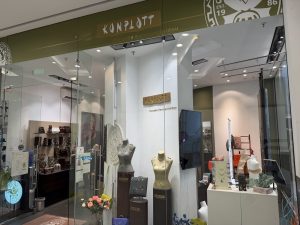 Konplott Miranda Konstantinidou shopfront at Belval Plaza, an olive green fascia with the Konplott wordmark and "Est. 1986" roundel, window display of necklaces and earrings on mannequin busts, bags and scarves on a ladder display, and a bunch of pink roses near the entrance.