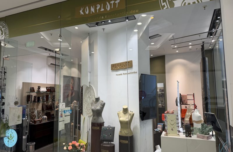 Konplott Miranda Konstantinidou shopfront at Belval Plaza, an olive green fascia with the Konplott wordmark and "Est. 1986" roundel, window display of necklaces and earrings on mannequin busts, bags and scarves on a ladder display, and a bunch of pink roses near the entrance.