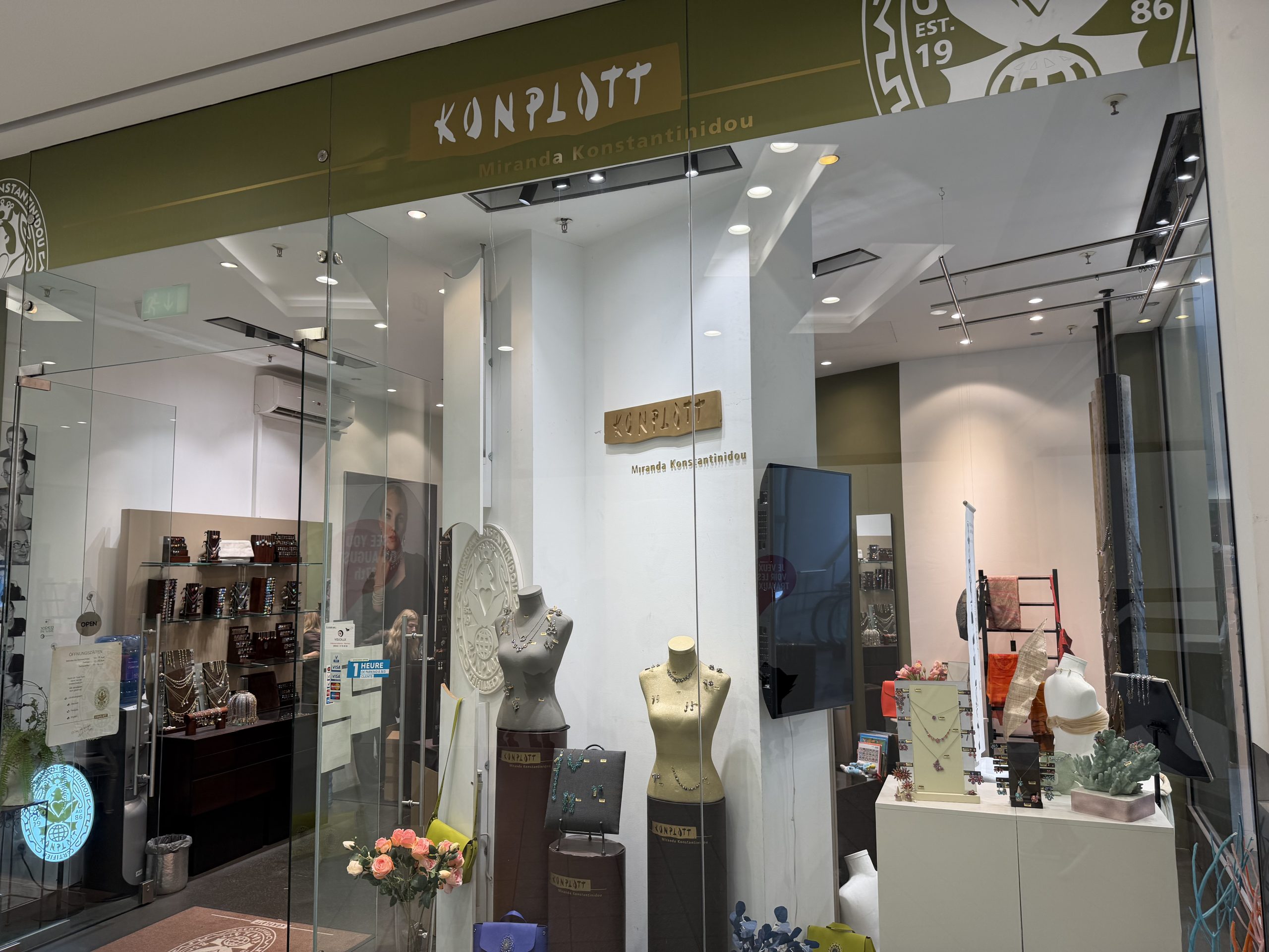 Konplott Miranda Konstantinidou shopfront at Belval Plaza, an olive green fascia with the Konplott wordmark and "Est. 1986" roundel, window display of necklaces and earrings on mannequin busts, bags and scarves on a ladder display, and a bunch of pink roses near the entrance.