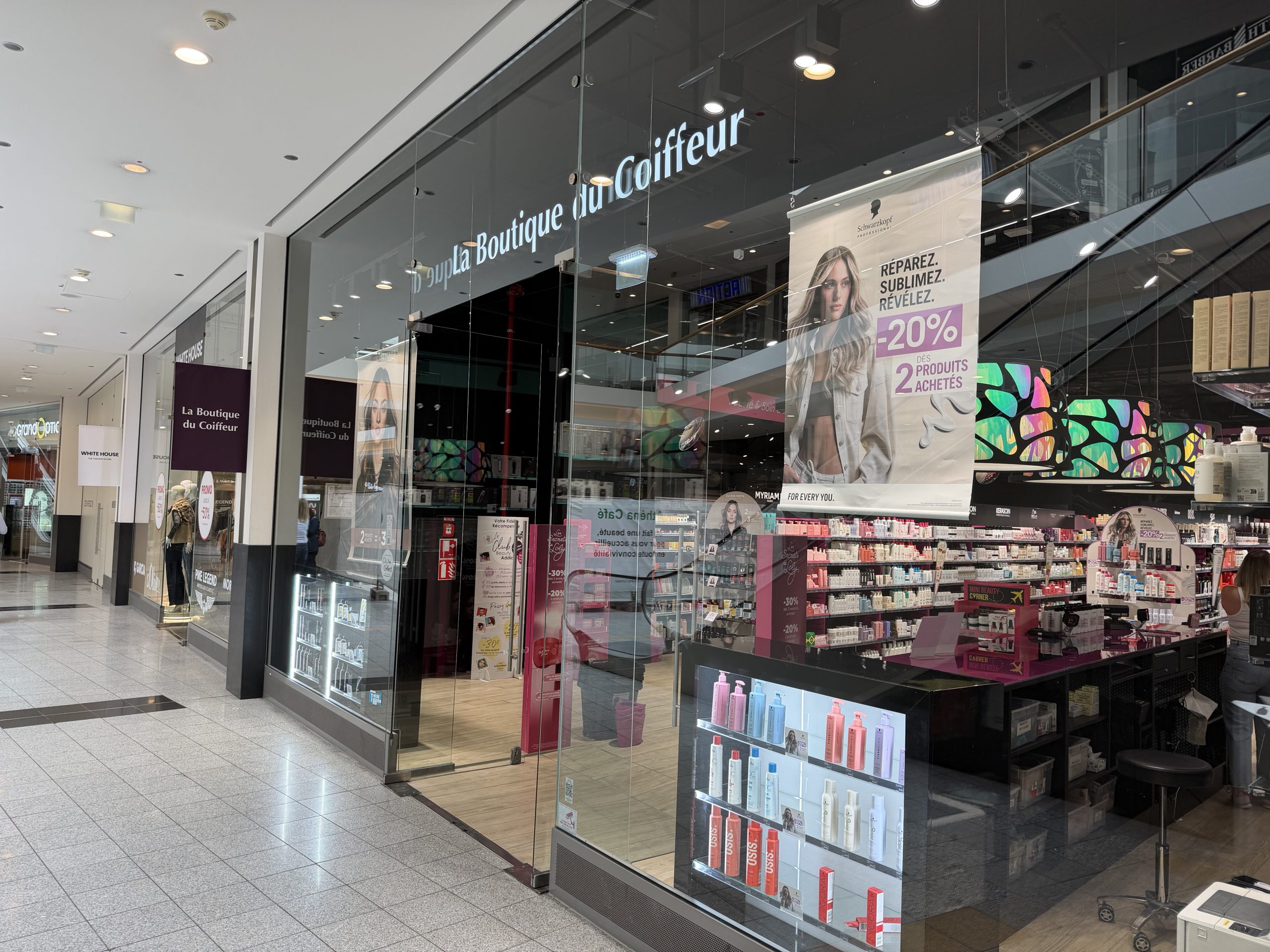 La Boutique du Coiffeur shopfront at Belval Plaza II, dark storefront with "La Boutique du Coiffeur" lettering, shelves of Schwarzkopf Professional haircare products, and a banner advertising twenty per cent off two products purchased together.