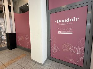The Le Boudoir by Belval Plaza door, a deep rose pink panel with white "le Boudoir" script lettering, "by BELVAL PLAZA" underneath, "Créateur de style" in a handwritten font, a QR code and "PERSONAL SHOPPER, Service sur rendez-vous" text, and gold line-drawn floral illustrations across the lower half of the door and the wall beside it.