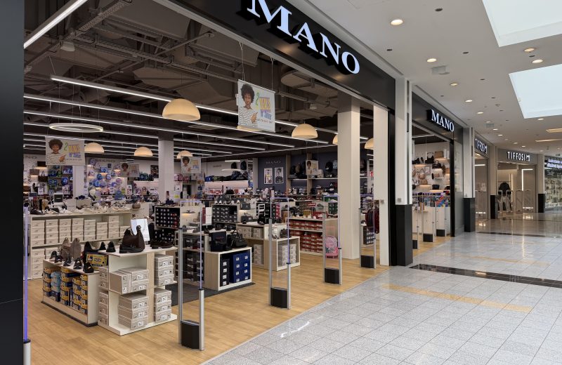 Mano shoe and bag shop at Belval Plaza, dark storefront with white "Mano" lettering, shelves of shoeboxes and handbags under pendant lighting, and a hanging sign advertising two deals for summer up to fifty per cent off.