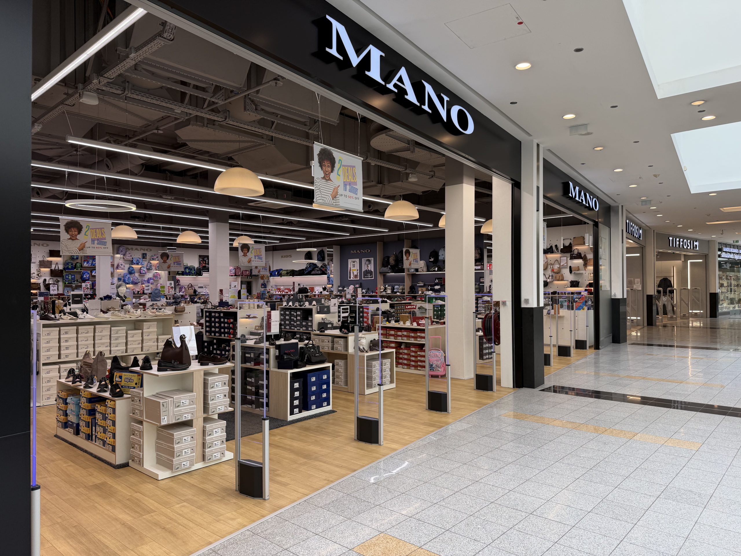Mano shoe and bag shop at Belval Plaza, dark storefront with white "Mano" lettering, shelves of shoeboxes and handbags under pendant lighting, and a hanging sign advertising two deals for summer up to fifty per cent off.