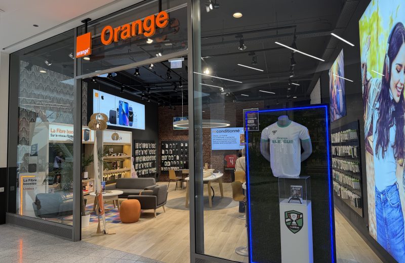 The Orange shopfront in Belval Plaza, with the orange Orange sign above the entrance, phone displays and seating inside, and a football shirt in a lit display case in the window.