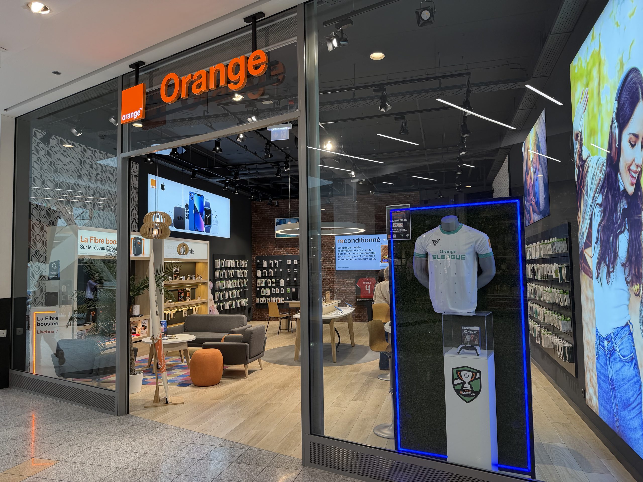 The Orange shopfront in Belval Plaza, with the orange Orange sign above the entrance, phone displays and seating inside, and a football shirt in a lit display case in the window.