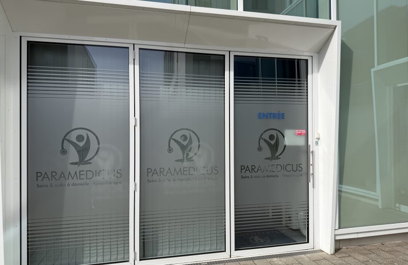 The entrance to Paramedicus at 4 rue John Lennon in Belvaux, with the company name and the words soins et aides à domicile, kinésithérapie etched on the glass.