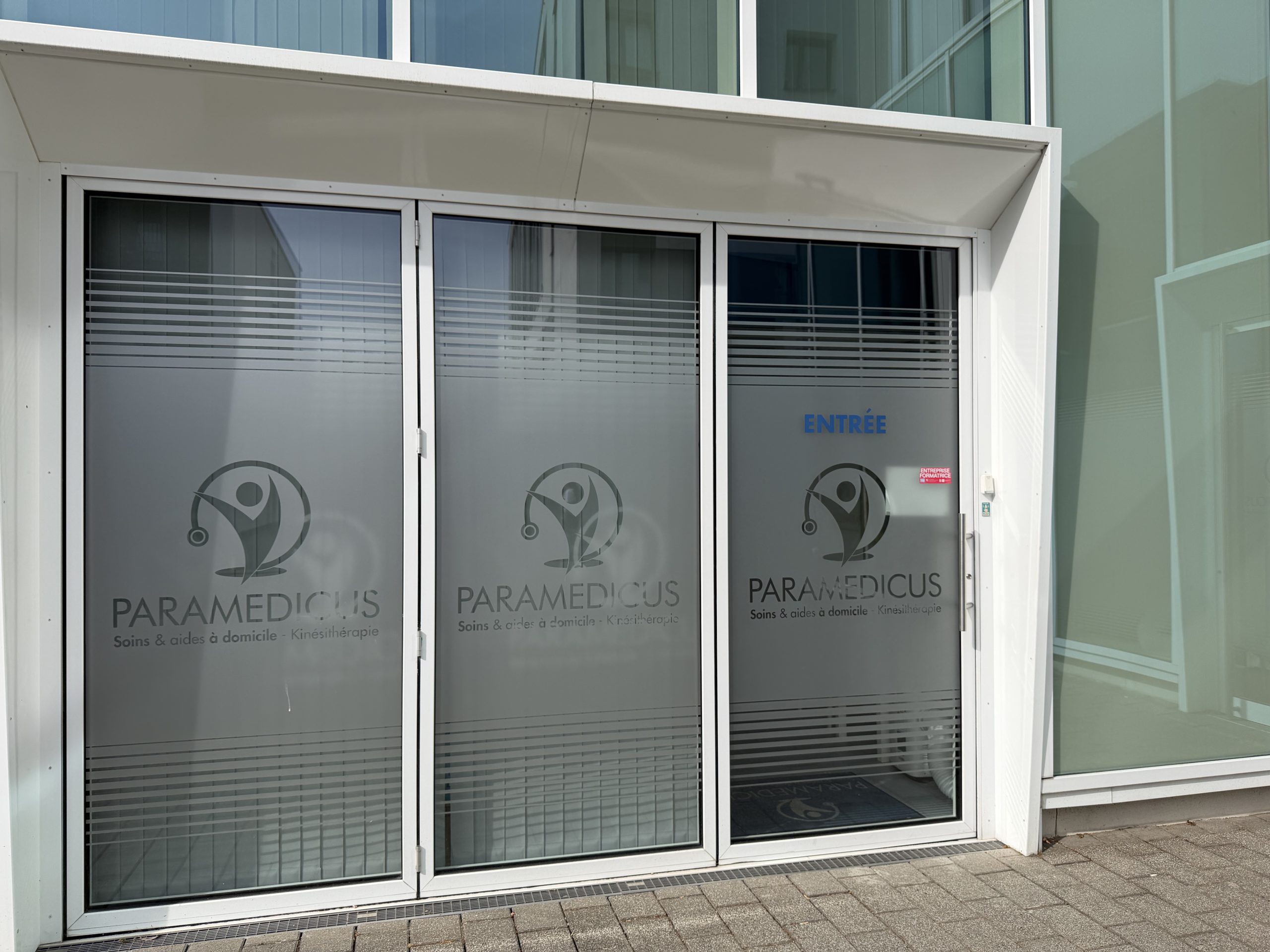 The entrance to Paramedicus at 4 rue John Lennon in Belvaux, with the company name and the words soins et aides à domicile, kinésithérapie etched on the glass.