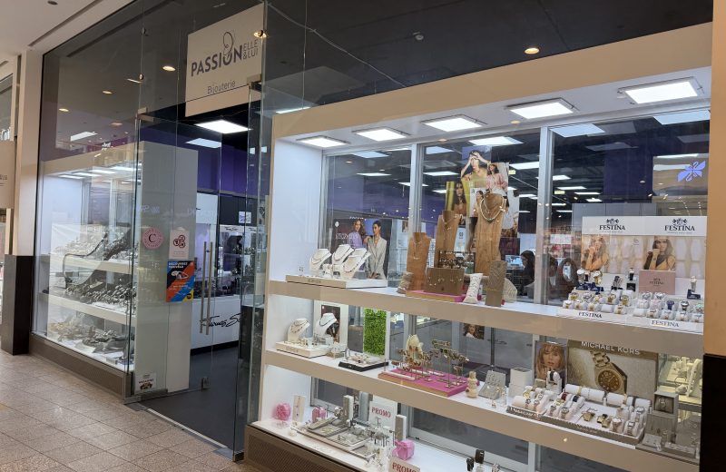 Passion'Elle & Lui bijouterie shopfront at Belval Plaza, glass storefront reading "Passion Elle & Lui, Bijouterie," window display of watches and necklaces on white risers with Festina and Michael Kors branding, and a colourful "Découvrez Taxivélo" poster near the door.