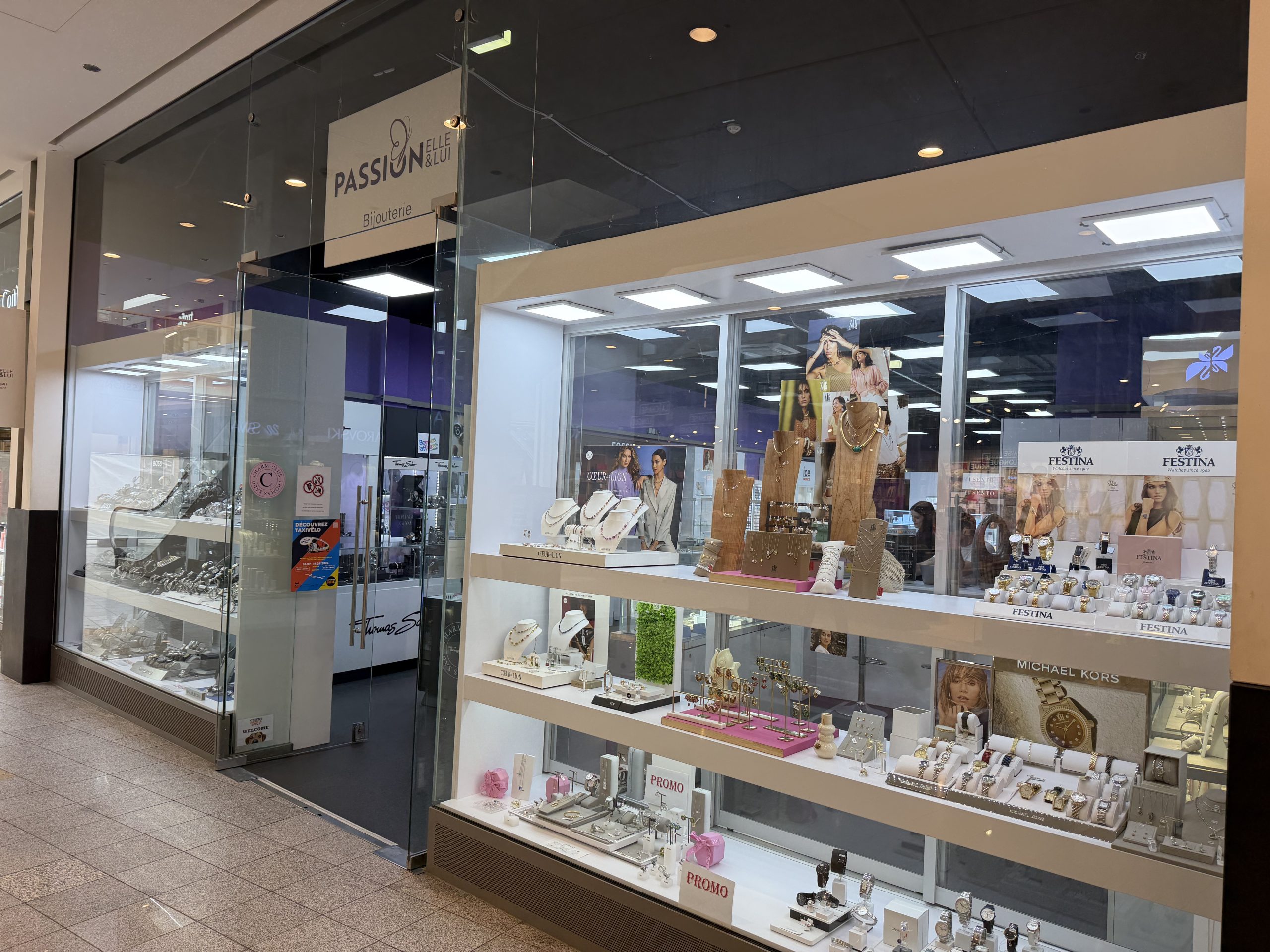 Passion'Elle & Lui bijouterie shopfront at Belval Plaza, glass storefront reading "Passion Elle & Lui, Bijouterie," window display of watches and necklaces on white risers with Festina and Michael Kors branding, and a colourful "Découvrez Taxivélo" poster near the door.