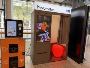 A Photomaton photo booth at Belval Plaza, grey cabin with a curtained entrance and a round red stool inside, screens displaying "Payment accepted," ID photo compliance flags for the Netherlands, Belgium and Germany, and a "no cash" notice, with an orange ME Group photo print kiosk standing beside it.