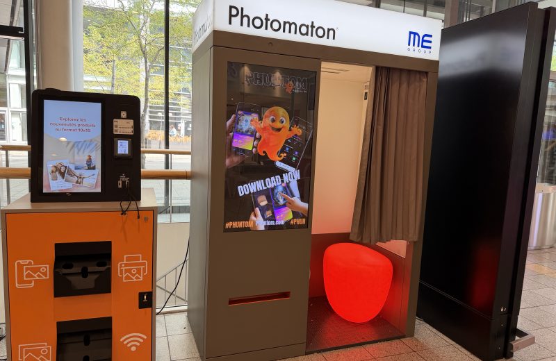 A Photomaton photo booth at Belval Plaza, grey cabin with a curtained entrance and a round red stool inside, screens displaying "Payment accepted," ID photo compliance flags for the Netherlands, Belgium and Germany, and a "no cash" notice, with an orange ME Group photo print kiosk standing beside it.