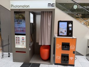 A second Photomaton photo booth at Belval Plaza, near an escalator, its screen displaying the "Phuntom" app branding with a ghost mascot and a "download now" prompt, red interior lighting visible through the curtained entrance, with the same style of orange ME Group print kiosk alongside it.