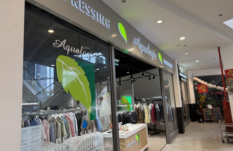 Aqualogia pressing shopfront at Belval Plaza II, grey storefront with backlit "Pressing" and "Aqualogia" lettering beside a green leaf logo, racks of pressed shirts and garments in plastic covers visible through the glass, and a counter with a card payment terminal near the entrance.
