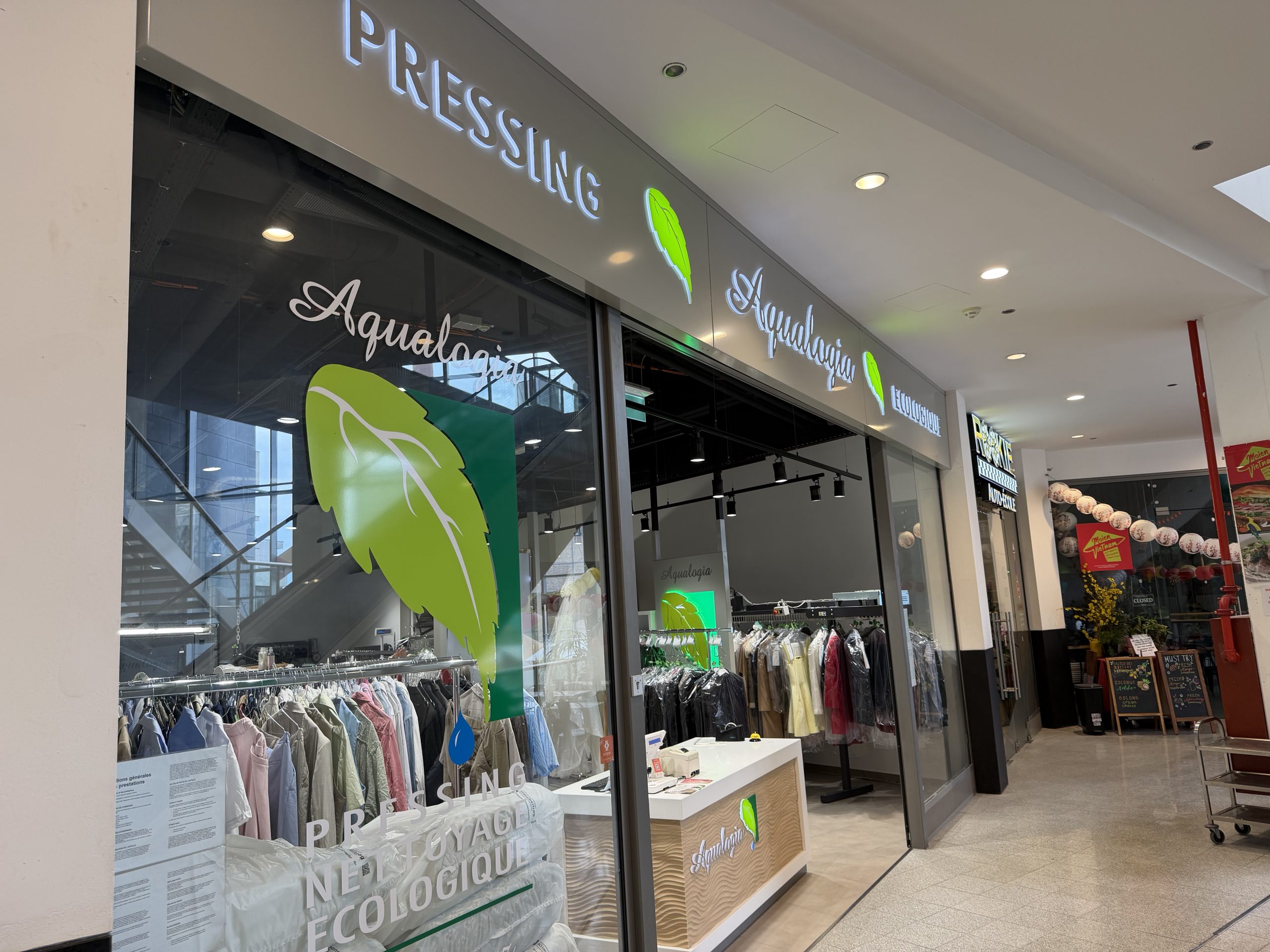Aqualogia pressing shopfront at Belval Plaza II, grey storefront with backlit "Pressing" and "Aqualogia" lettering beside a green leaf logo, racks of pressed shirts and garments in plastic covers visible through the glass, and a counter with a card payment terminal near the entrance.