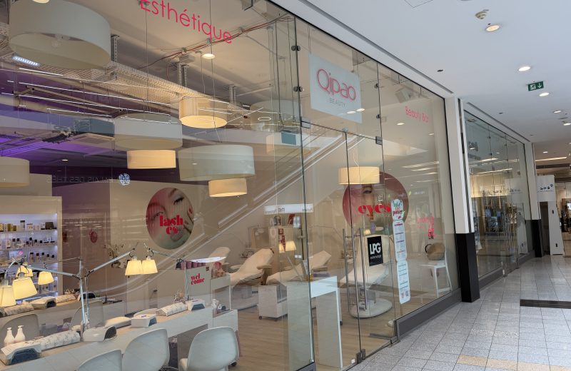 Qipao Beauty shopfront at Belval Plaza II, glass frontage with the Qipao Beauty wordmark and "Esthétique" in pink neon lettering, a nail and lash bar with pendant lighting and treatment chairs visible through the glass, and circular window graphics for lash and eye treatments.