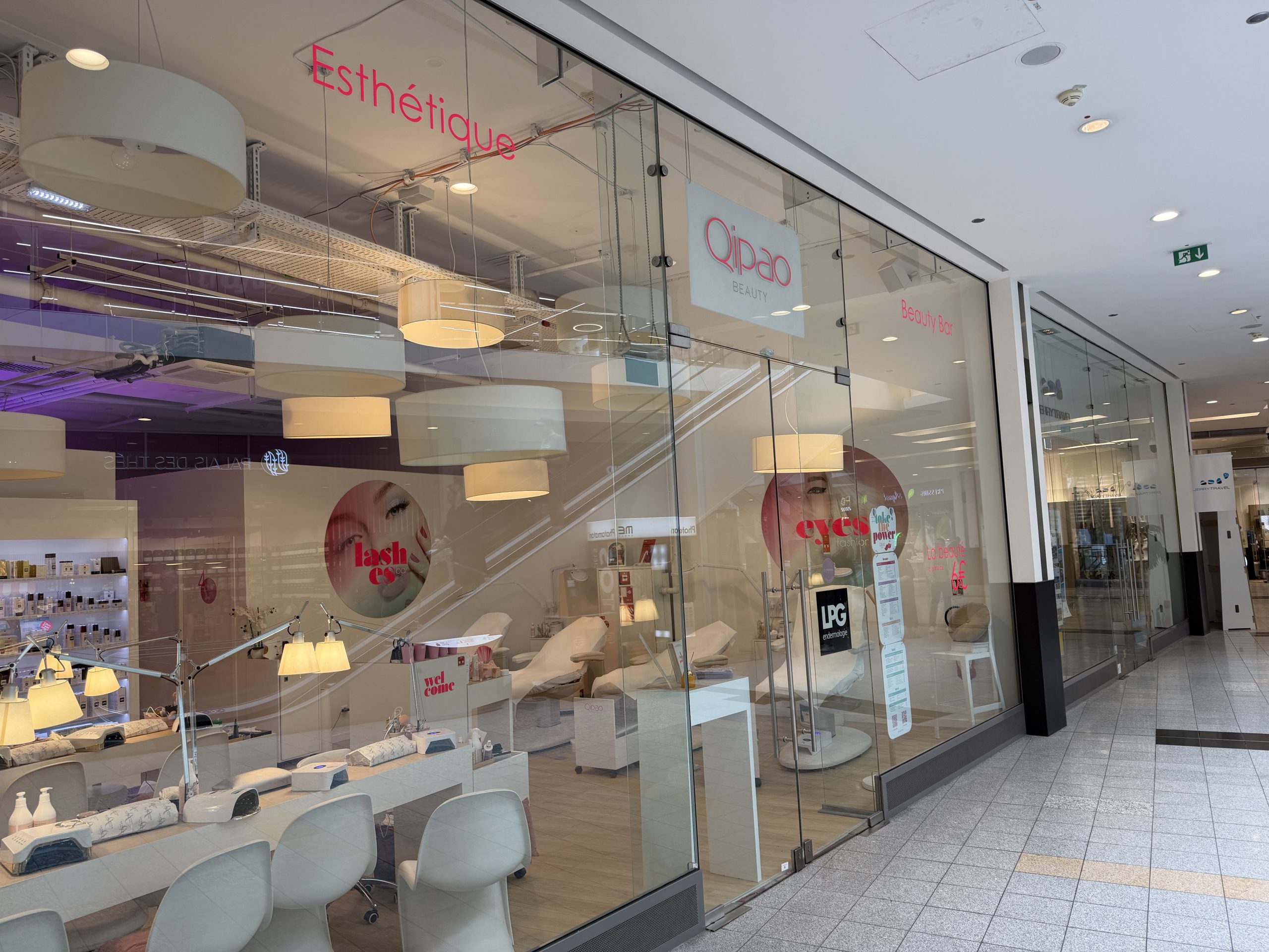 Qipao Beauty shopfront at Belval Plaza II, glass frontage with the Qipao Beauty wordmark and "Esthétique" in pink neon lettering, a nail and lash bar with pendant lighting and treatment chairs visible through the glass, and circular window graphics for lash and eye treatments.