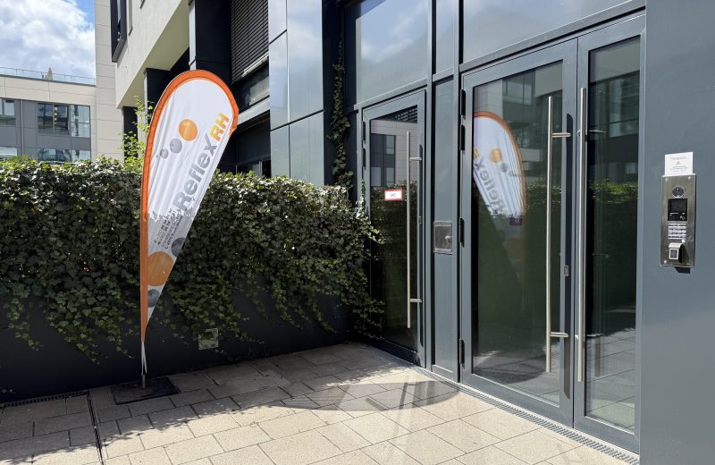 The entrance to the Belval Business Center on boulevard du Jazz, with an orange and white Reflex RH teardrop flag standing beside an ivy-covered wall, glass doors with a camera intercom and fingerprint reader, and a small Reflex RH nameplate giving the bell number.
