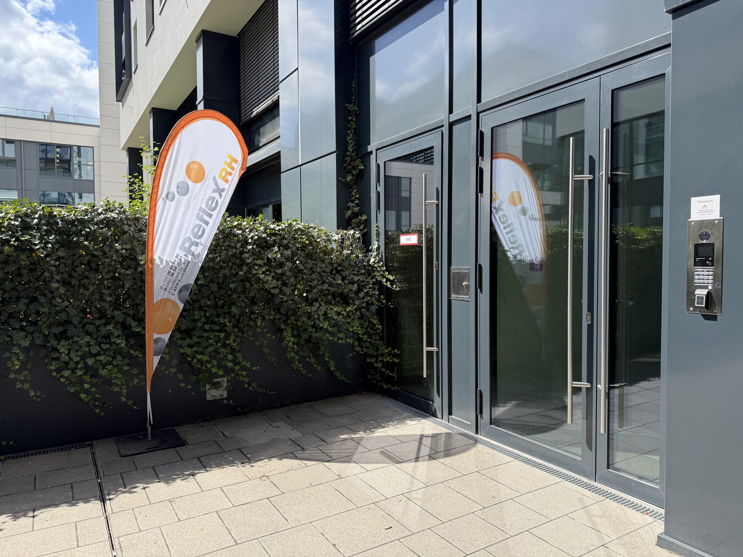 The entrance to the Belval Business Center on boulevard du Jazz, with an orange and white Reflex RH teardrop flag standing beside an ivy-covered wall, glass doors with a camera intercom and fingerprint reader, and a small Reflex RH nameplate giving the bell number.