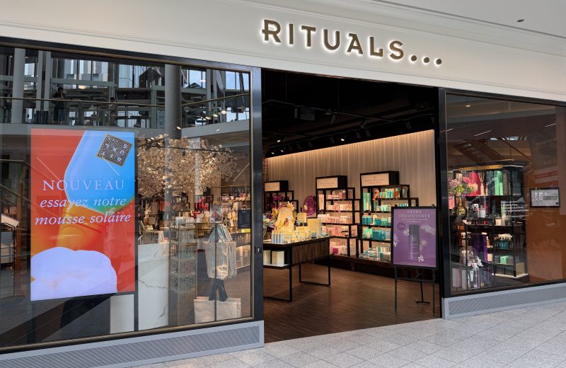 The Rituals shopfront in Belval Plaza, with the gold RITUALS sign above the entrance, a white blossom display near the door and dark wood shelving of body care and home fragrance products inside.