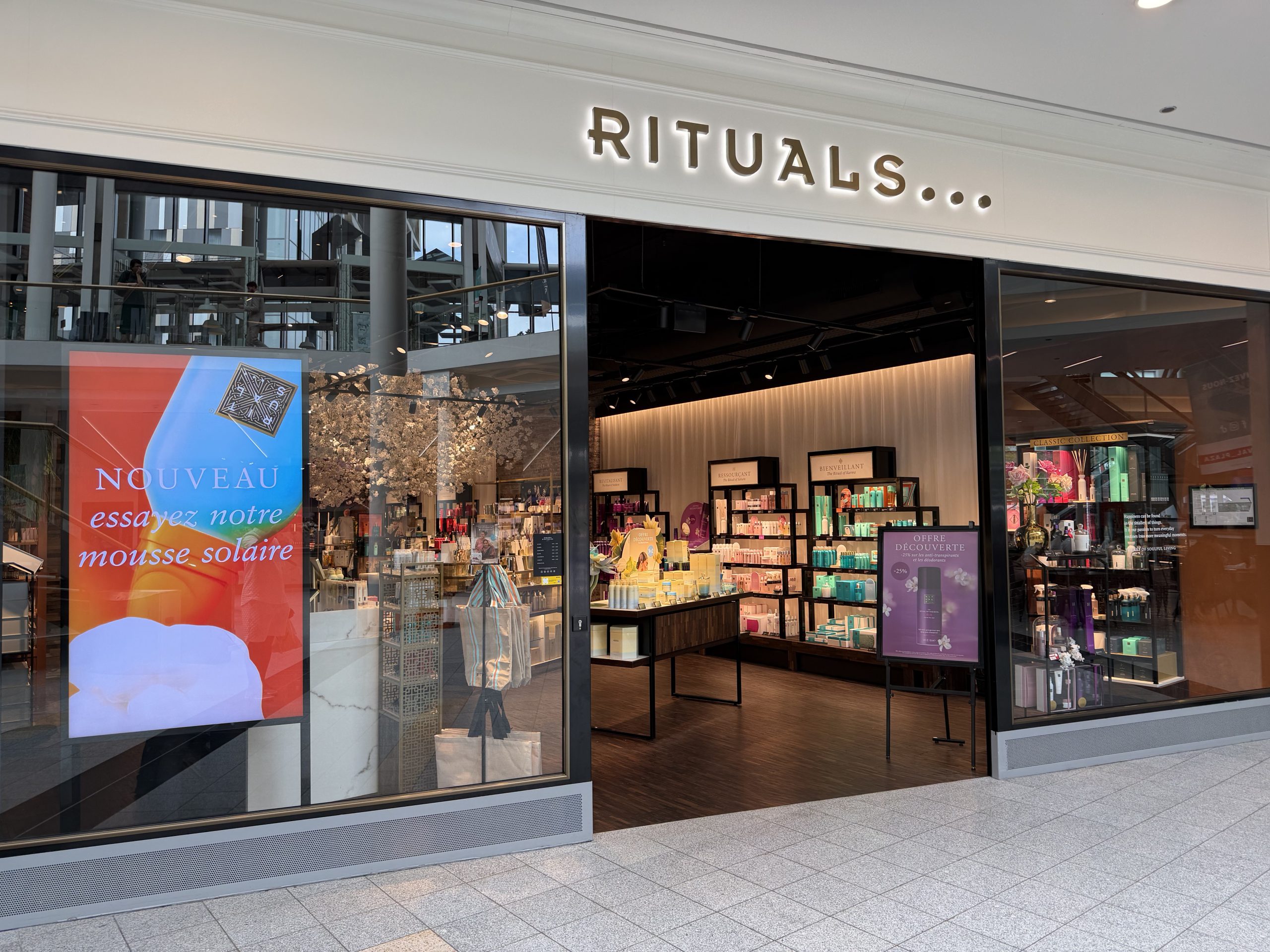 The Rituals shopfront in Belval Plaza, with the gold RITUALS sign above the entrance, a white blossom display near the door and dark wood shelving of body care and home fragrance products inside.
