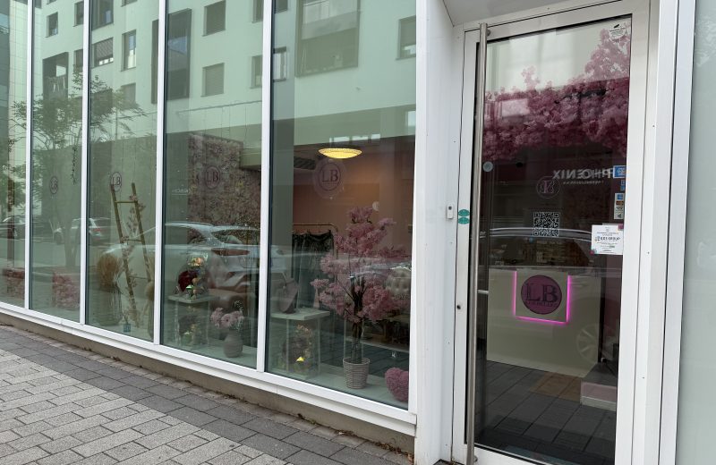 The shopfront of Les Belles in Belval, a beauty salon with a pink neon LB logo on the glass door, card payment and contactless stickers, and a window display of pink artificial cherry blossom, a flower wall, a cream armchair, jewellery on stands and a clothing rail inside.